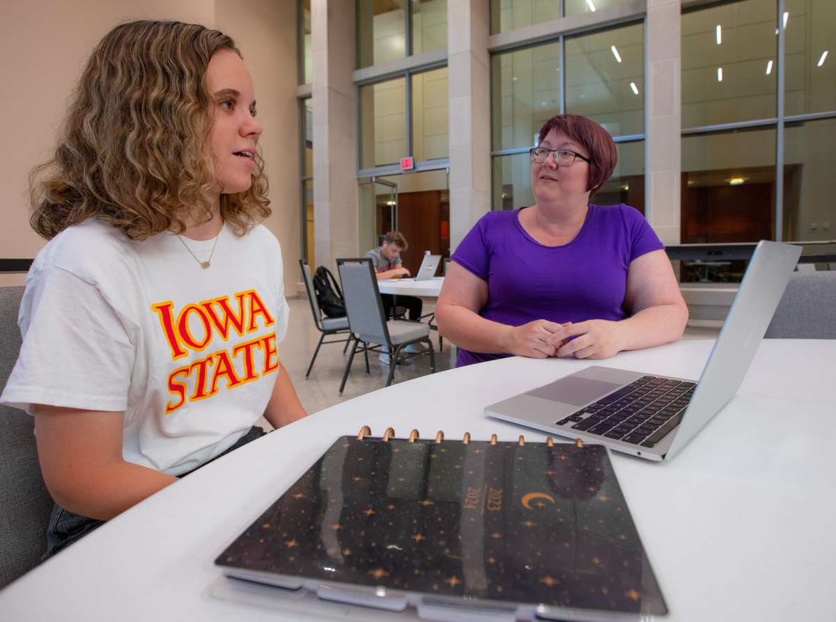 A woman in an Iowa State t-shirt and a woman in a purple t-shirt sit at a table with a laptop and notebook on it.